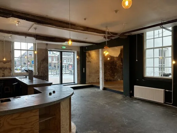 Industrieel ingerichte horecaruimte met houten bar, betonnen vloer en uitzicht op het Sint-Agathaplein.